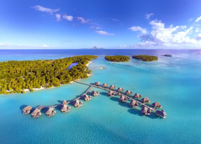 Vue aérienne suites et villas sur pilotis, motu, lagon turquoise et île de Bora Bora au loin - Hôtel Le Taha'a by Pearl Resorts 5*****